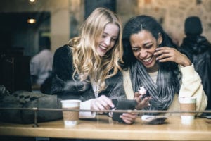Two multiethnic young female friends having fun, rejoicing memories using a smart phone while enjoying coffee and pastries in the local NYC coffee shop, natural light, taken from the street.